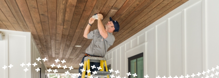 Technician standing on a ladder installing or repairing a ceiling fixture on a wood-paneled porch ceiling.