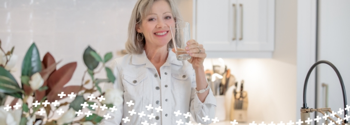 Smiling woman holding a glass of water in a bright modern kitchen with plants in the foreground.