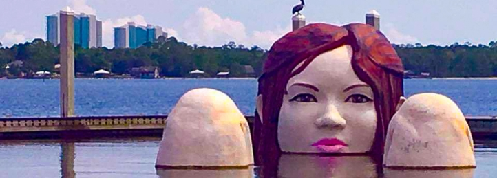 Large floating sculpture of a woman's face in the water with a lake and city skyline in the background.