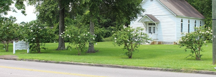 Small white church building with a front entrance and manicured lawn along a quiet roadside.