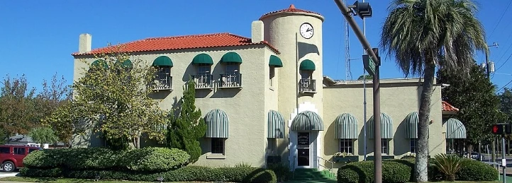 Mediterranean-style building with a clock tower, green awnings, and palm trees under a clear blue sky.