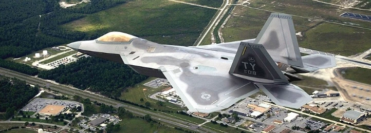 F-22 Raptor fighter jet flying over a military base and surrounding landscape from an aerial view.