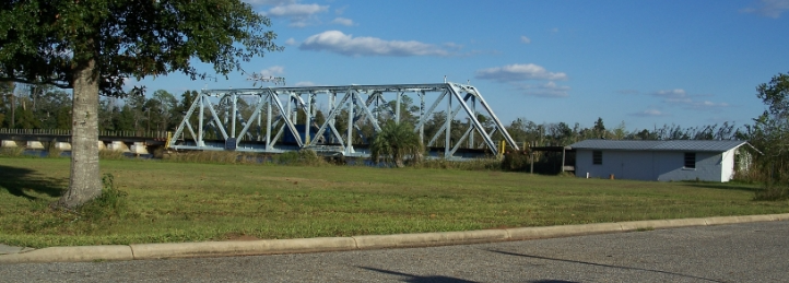Steel truss bridge spanning a river near a grassy roadside and small white building under a blue sky.
