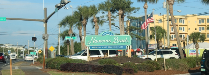 Navarre Beach welcome sign surrounded by palm trees, traffic, and beachfront buildings on a sunny day.